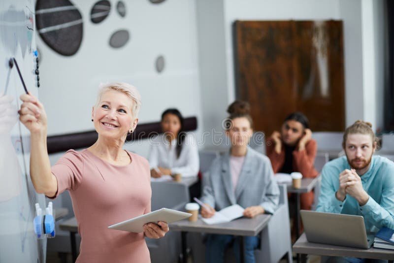 mature-female-teacher-waist-up-portrait-smiling-pointing-whiteboard-presenting-training-course-students-class-151369394